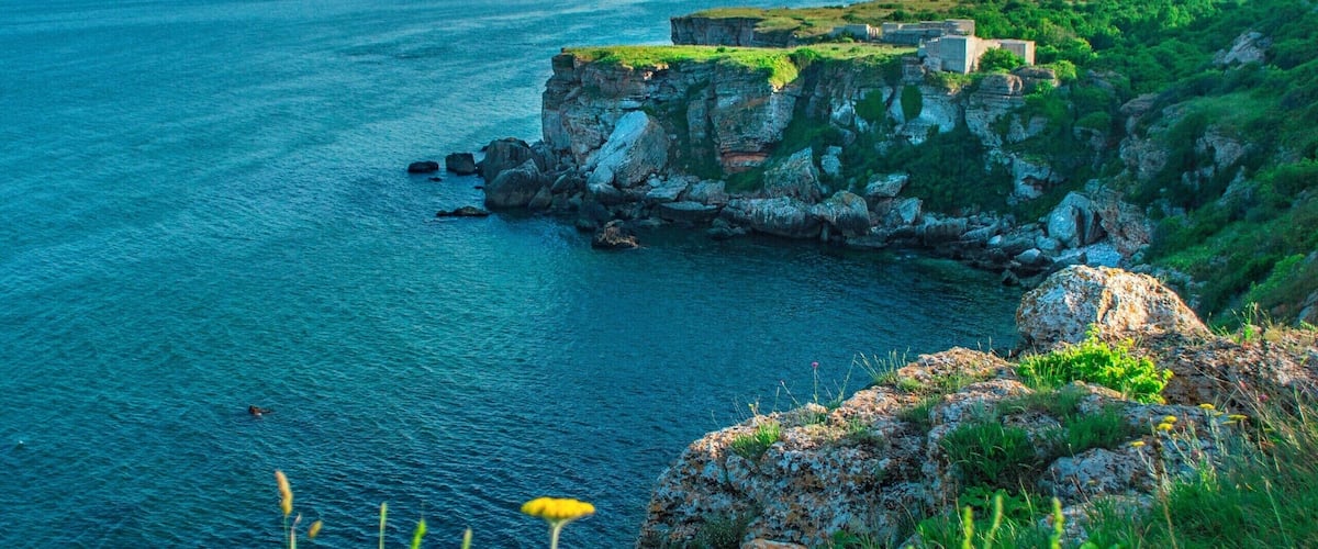 A coastal landscape with a rocky cliff, green vegetation, and a clear blue sea.