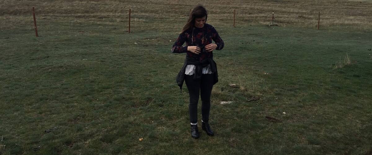 A person standing in a grassy field with a fence in the background and mountains in the distance.