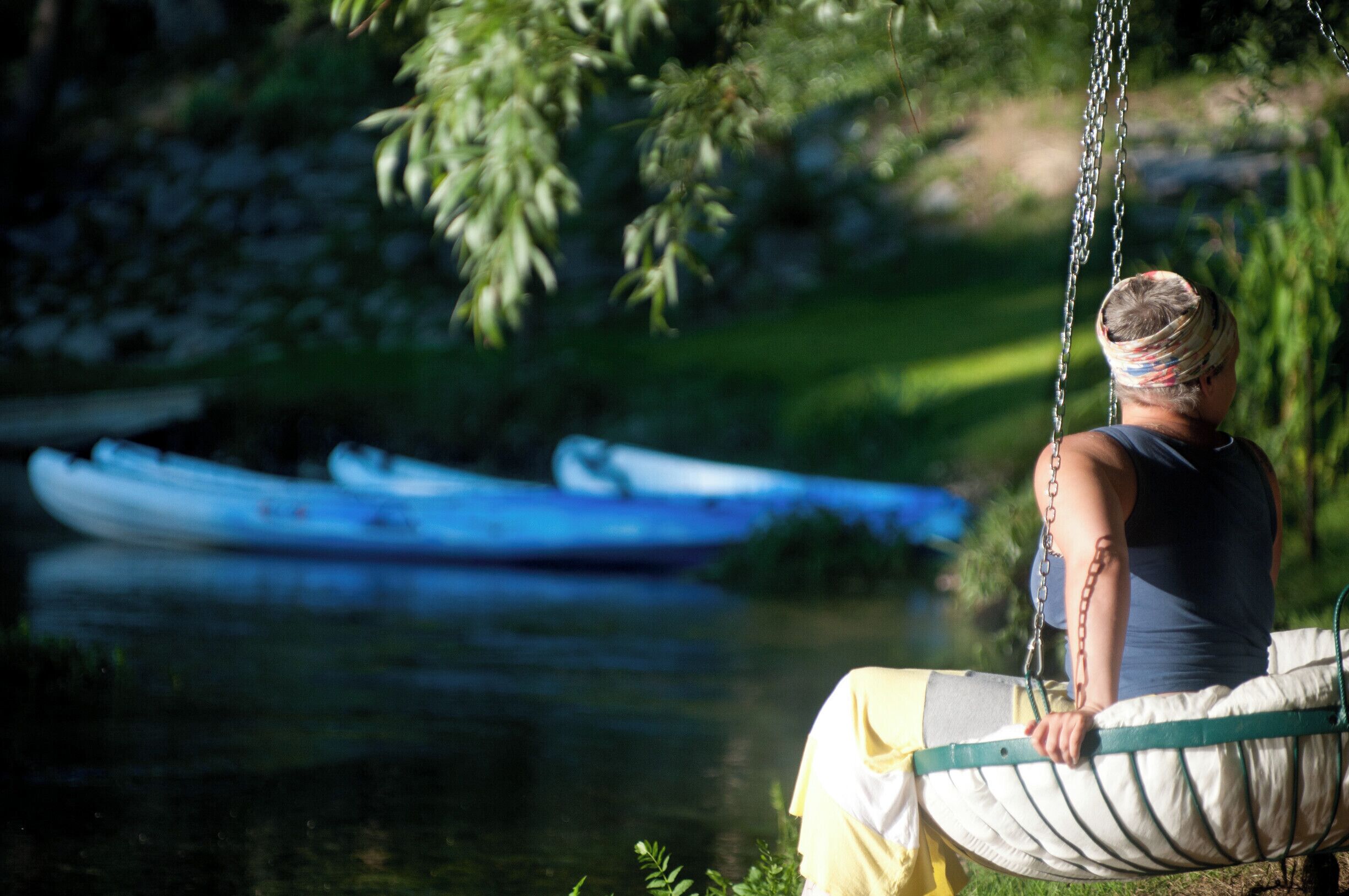A person sitting on a hammock by a calm river with boats in the background.