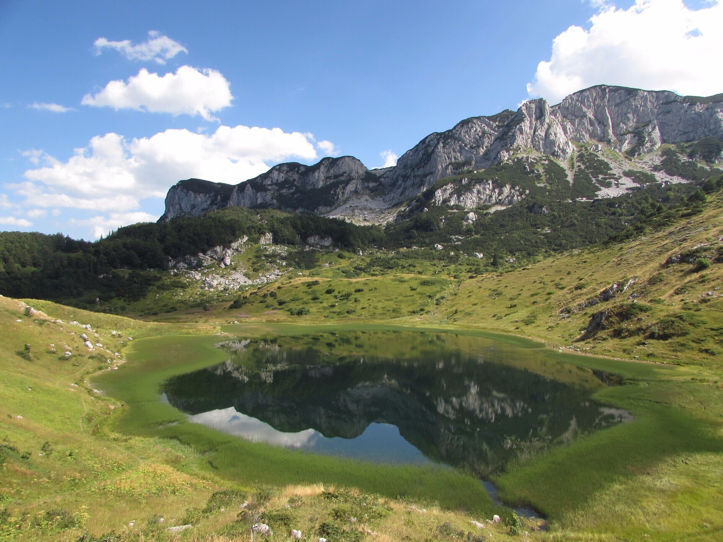 A mountain landscape with a lake reflecting the sky and surrounding greenery.