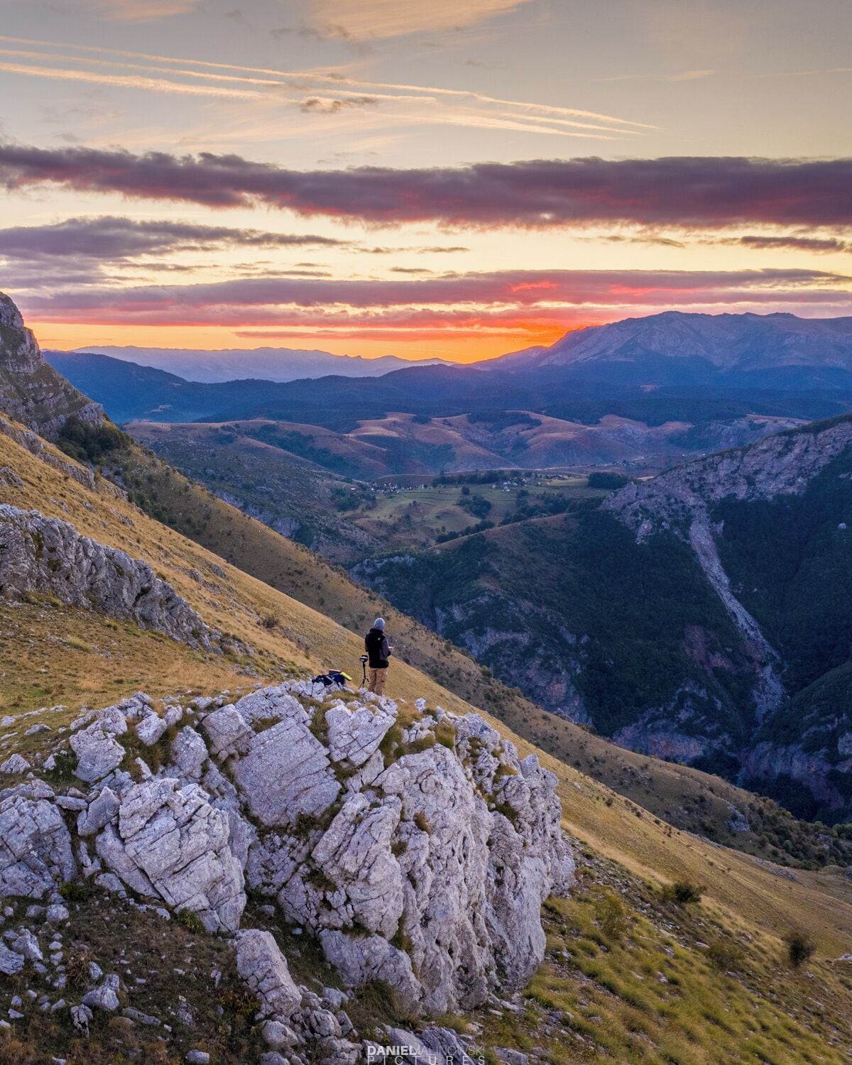 A hiker on a rocky outcrop overlooking a valley with distant mountains during sunset.