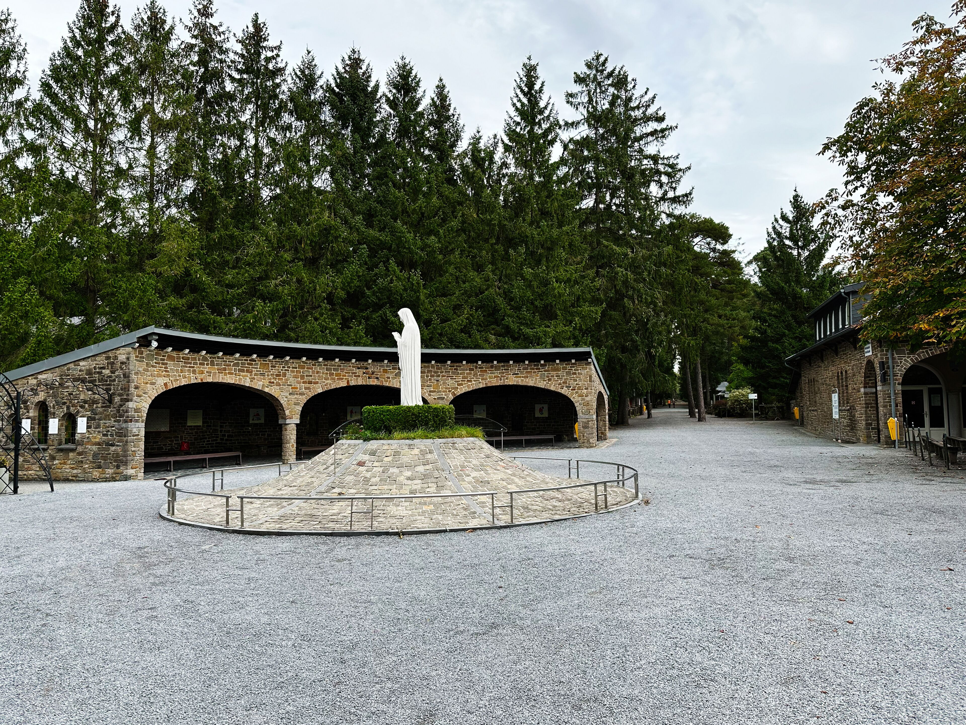 Statue of Our Lady in a circular stone pedestal surrounded by arches and trees at Belgian memorial site. Sanctuary Banneux.