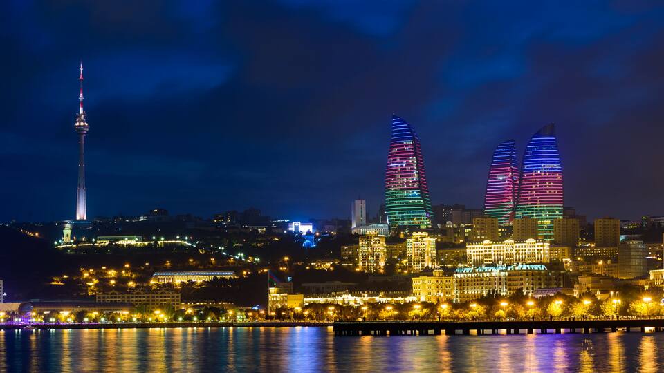 Une vue nocturne sur les gratte-ciel illuminés d’une ville, avec une tour imposante et une promenade au bord de l’eau où se reflètent les lumières.