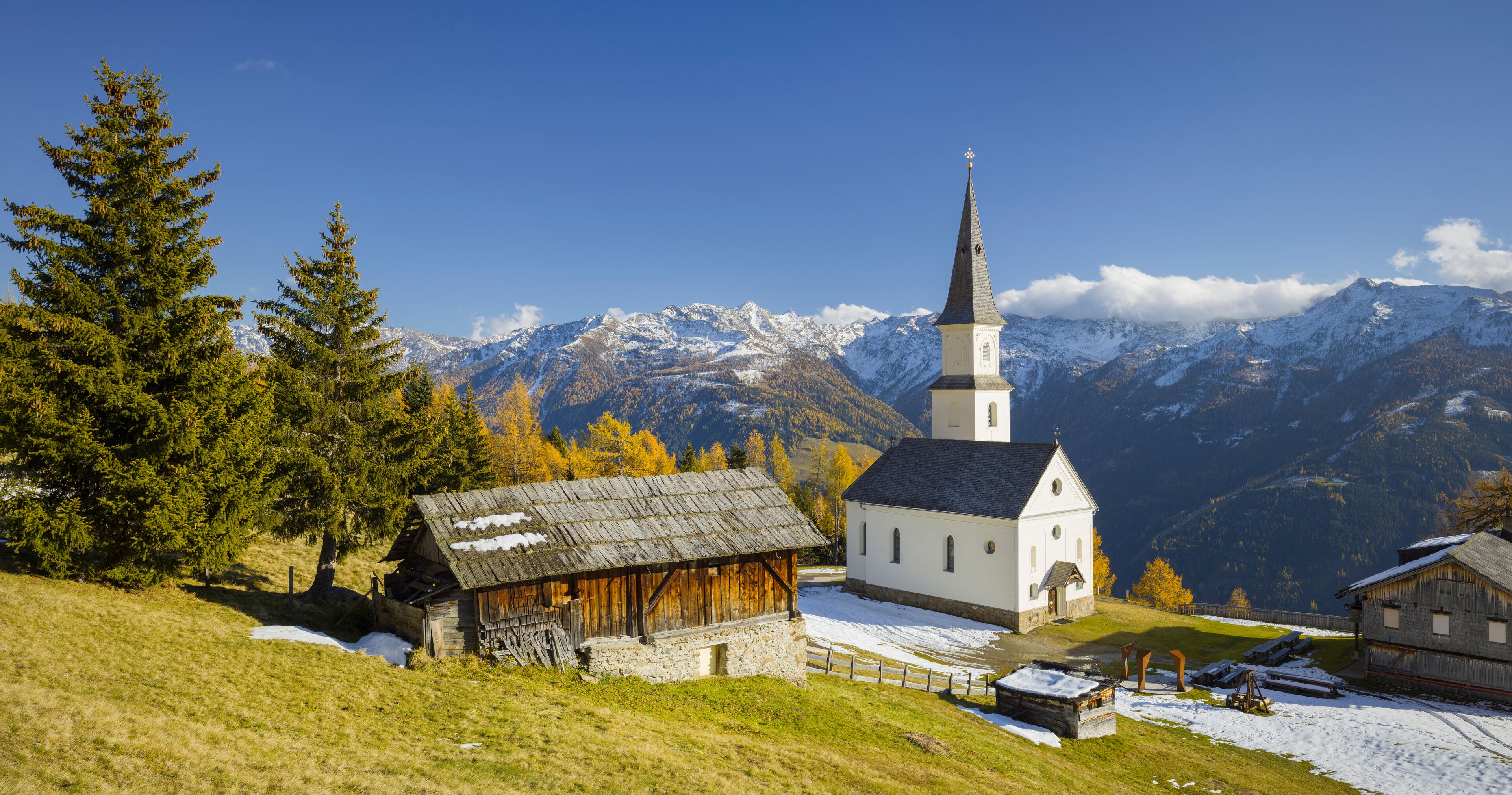 A church with a tall steeple, surrounded by snow-covered mountains and autumn trees.