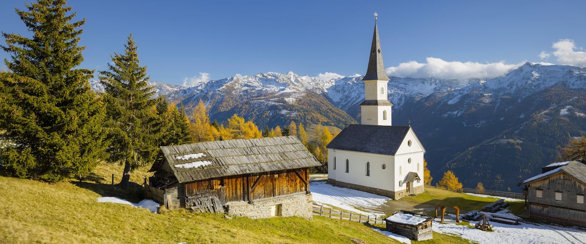 A church with a tall steeple, surrounded by snow-covered mountains and autumn trees.