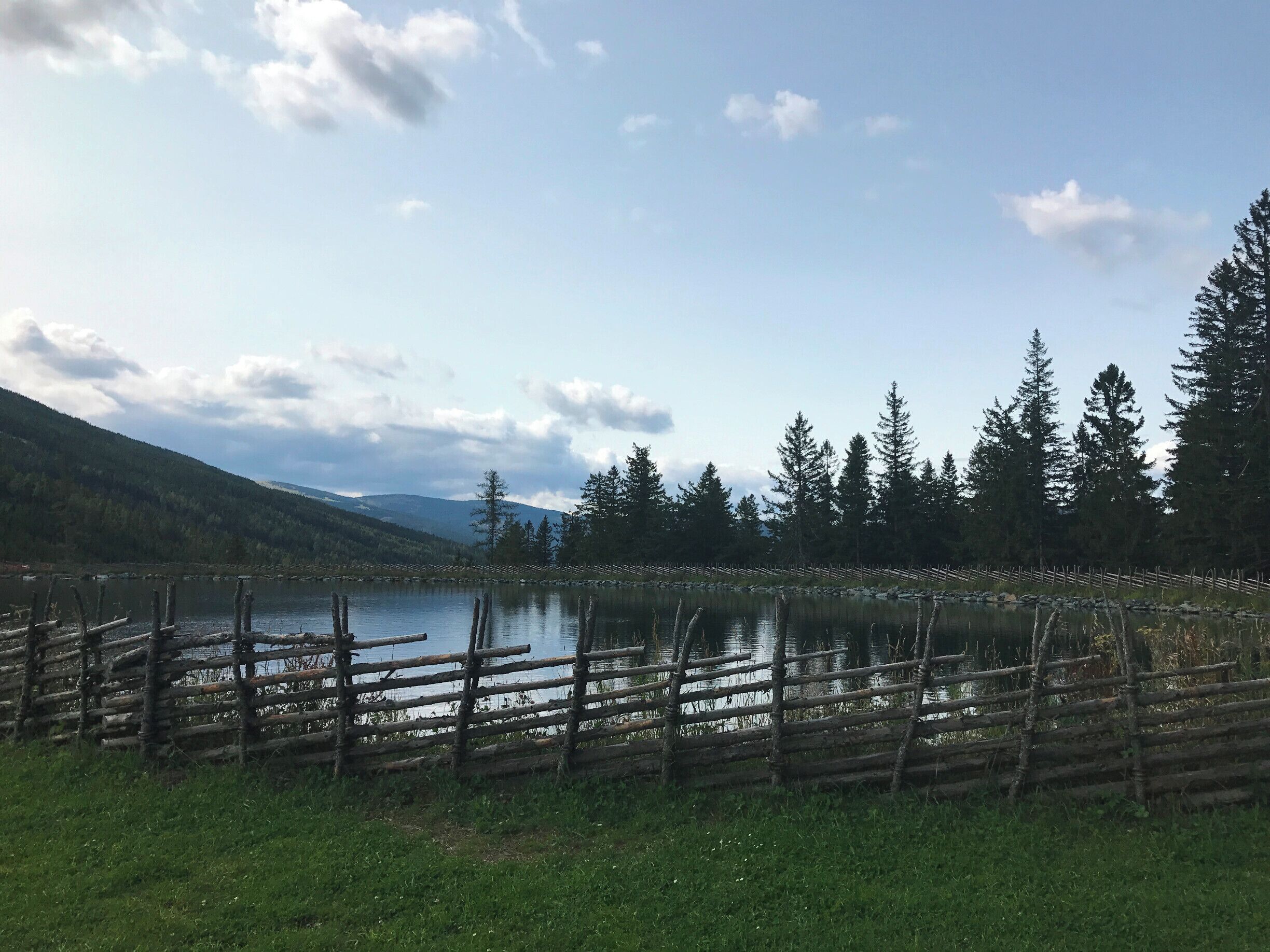 A lake surrounded by a wooden fence, trees, and mountains in the background.