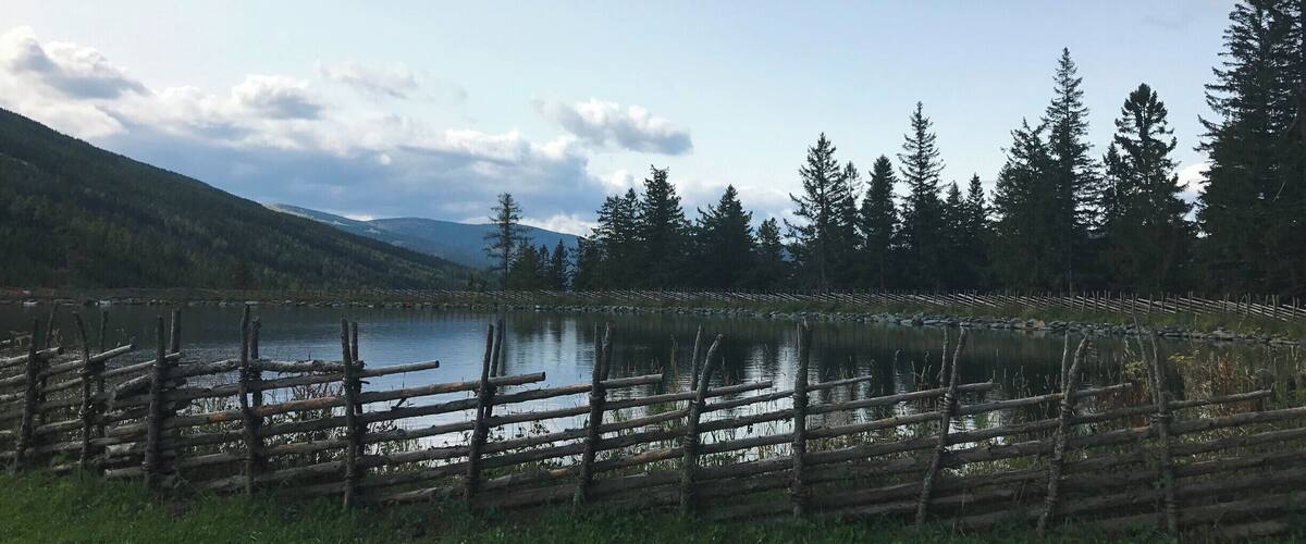 A lake surrounded by a wooden fence, trees, and mountains in the background.
