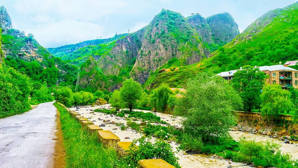 A narrow road winding through a lush valley with rocky cliffs and a river.