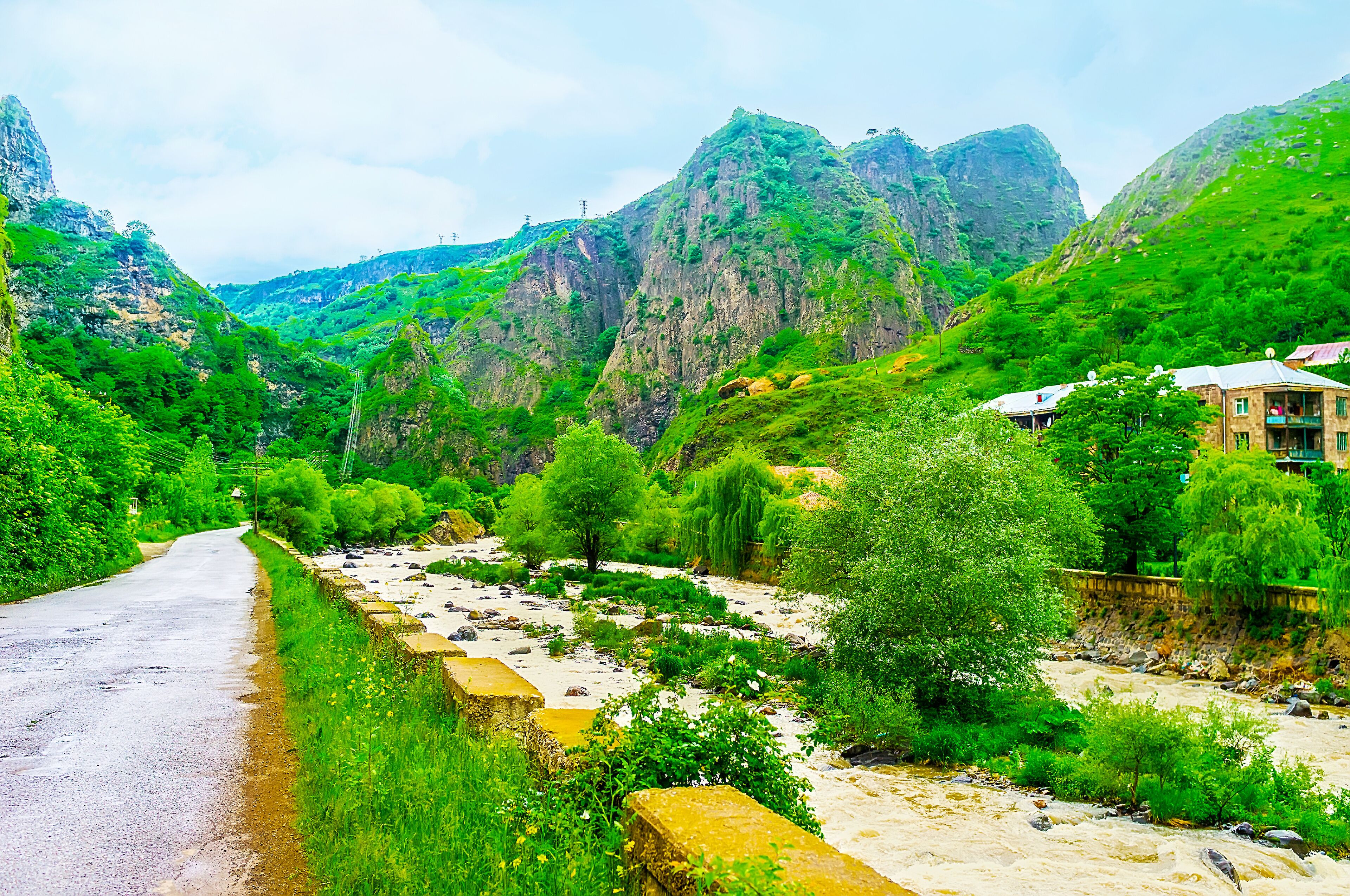 A narrow road winding through a lush valley with rocky cliffs and a river.