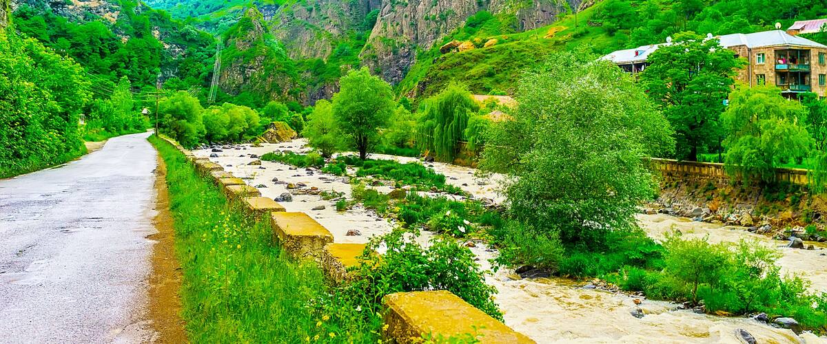 A narrow road winding through a lush valley with rocky cliffs and a river.