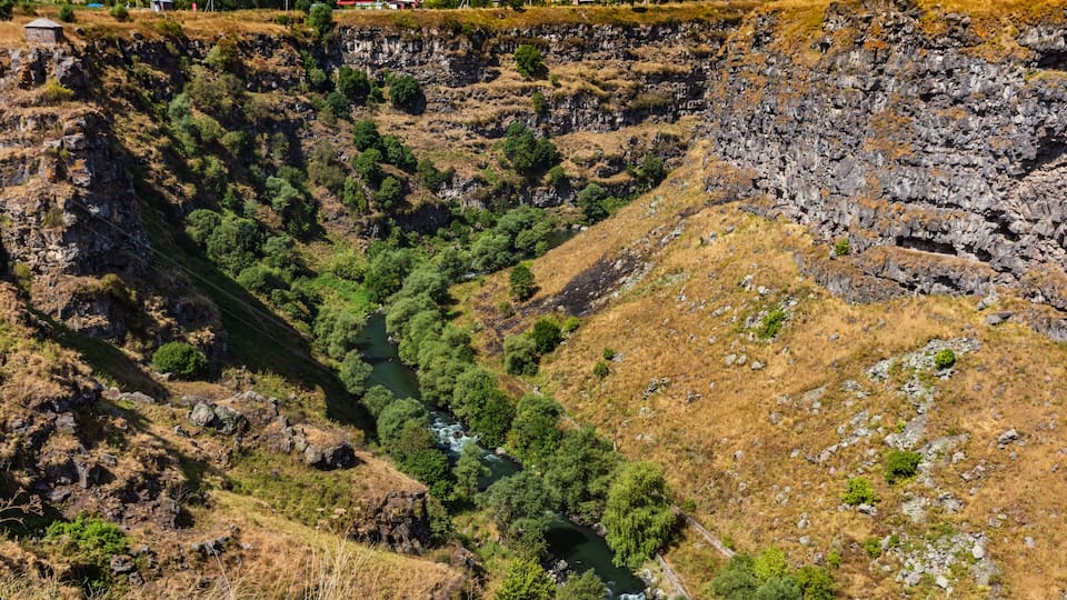 A river winding through a rocky gorge with sparse vegetation and a few buildings in the background.