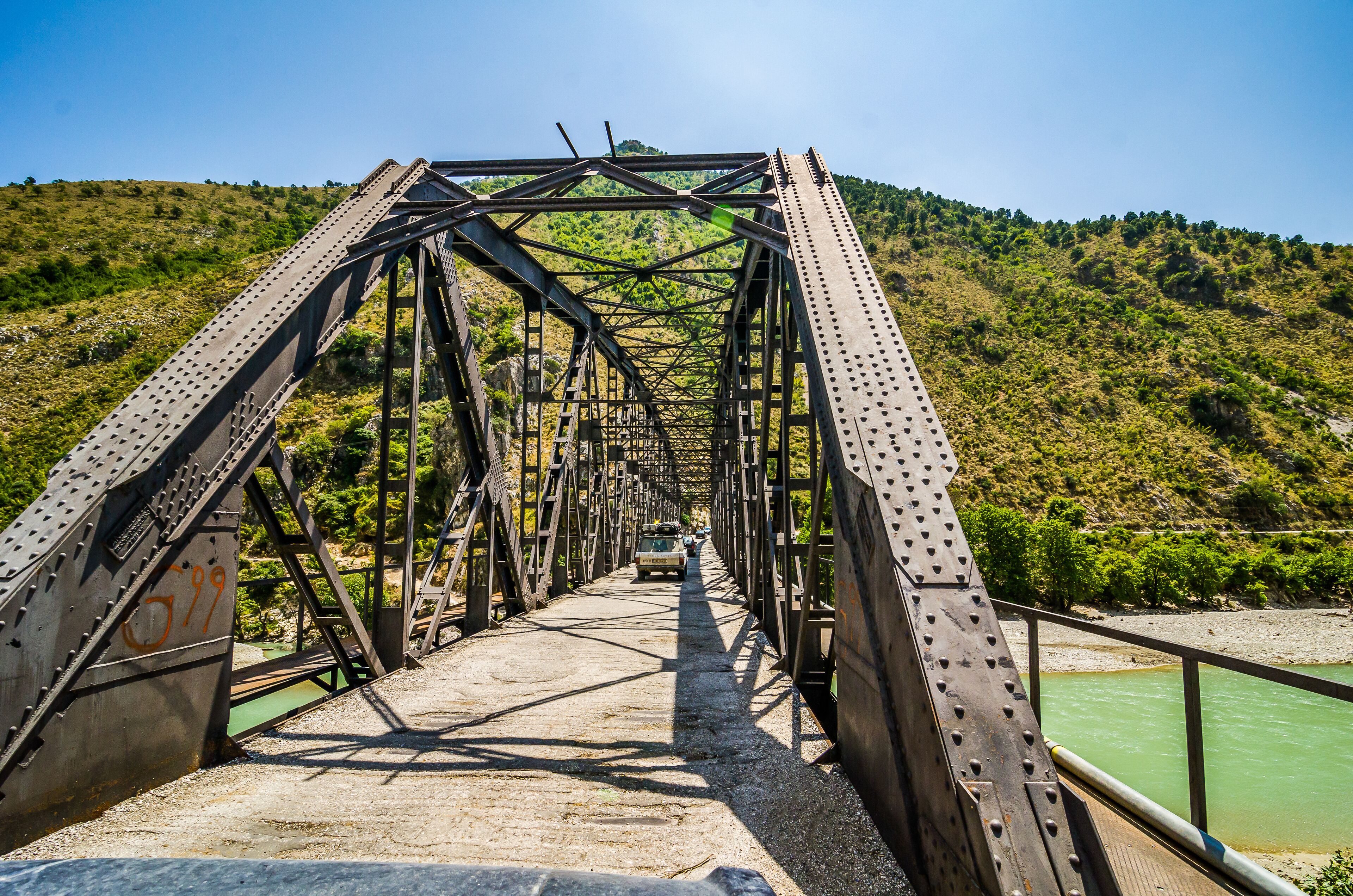 A metal bridge with a green river below, surrounded by hills.