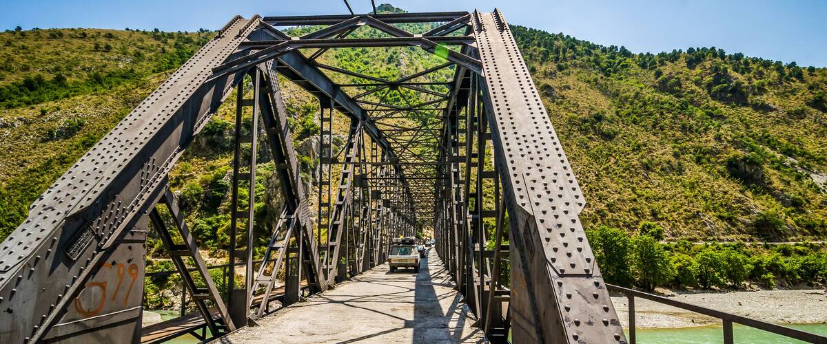 A metal bridge with a green river below, surrounded by hills.