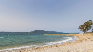 Uma praia de seixos com águas turquesa cristalinas, um molhe rochoso e montanhas ao longe.