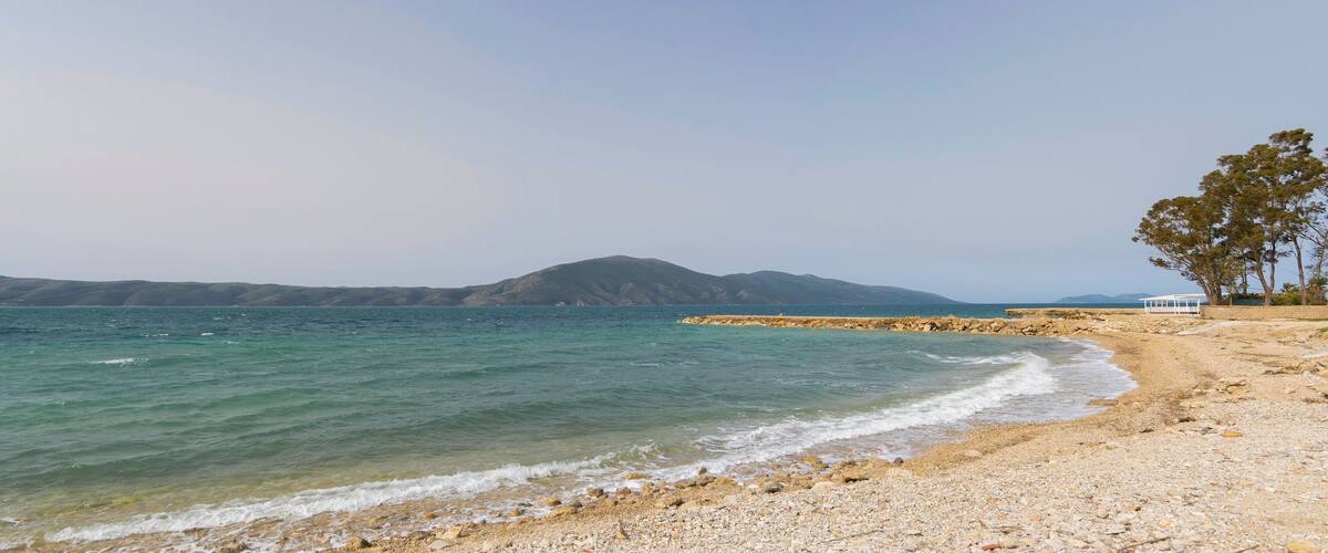 A pebble beach with clear turquoise water, a rocky breakwater, and distant mountains.