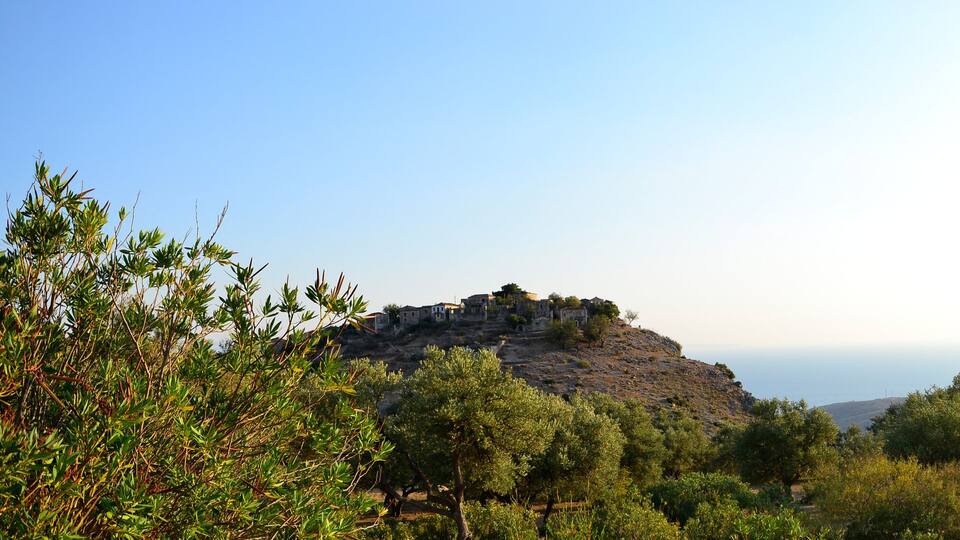 A mountainous landscape with olive trees, a small village on a hill, and ancient ruins.