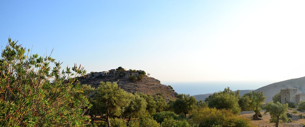 A mountainous landscape with olive trees, a small village on a hill, and ancient ruins.
