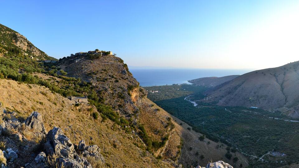 A mountainous landscape with a small building on a cliff, a river valley, and the sea in the distance.