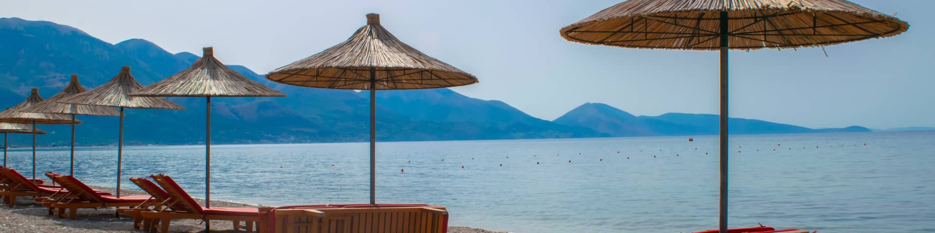 Beach umbrellas, and sun loungers on pebble beach. Qeparo village. Albania. Ionian Sea. Vacation sea concept. Summer sunny seaside landscape.