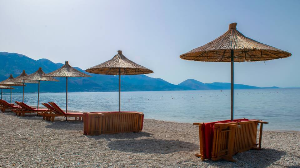 A beach with wooden lounge chairs and thatched umbrellas, a pebble beach, and mountains in the background.
