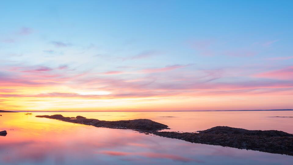A sunset over a calm body of water with rocky outcrops and a distant shoreline.