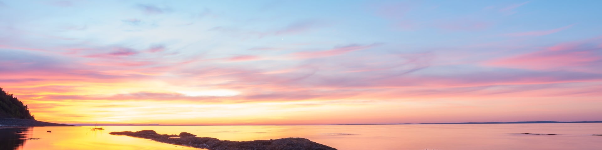 A sunset over a calm body of water with rocky outcrops and a distant shoreline.