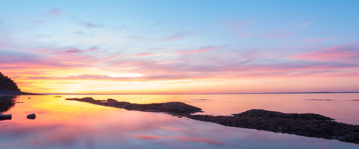 A sunset over a calm body of water with rocky outcrops and a distant shoreline.