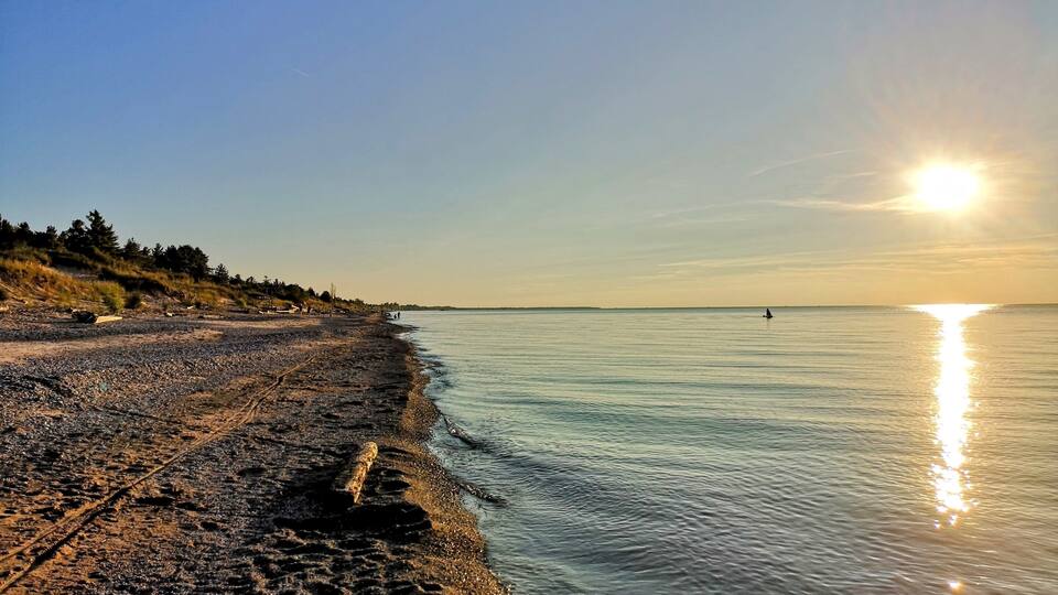 A pebbly beach with a clear sky and the sun setting over the water.