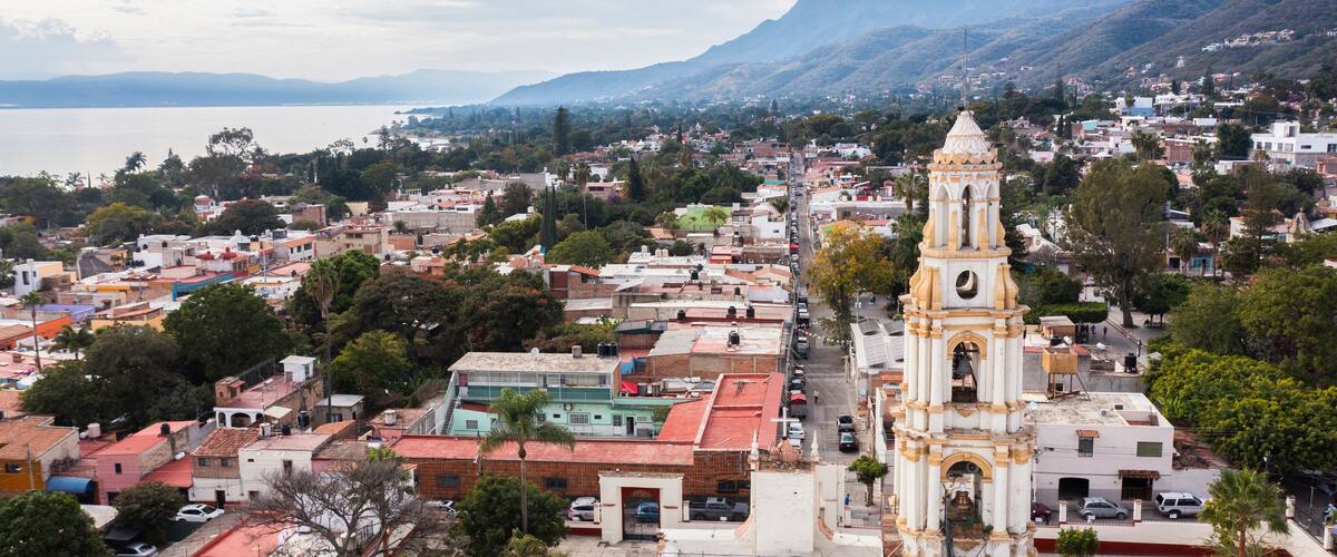 A coastal cityscape with a prominent white bell tower, lush greenery, and a mountain range in the background.