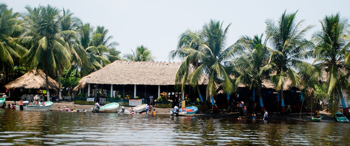 A tropical beach with thatched-roof huts, palm trees, and small boats on the water.