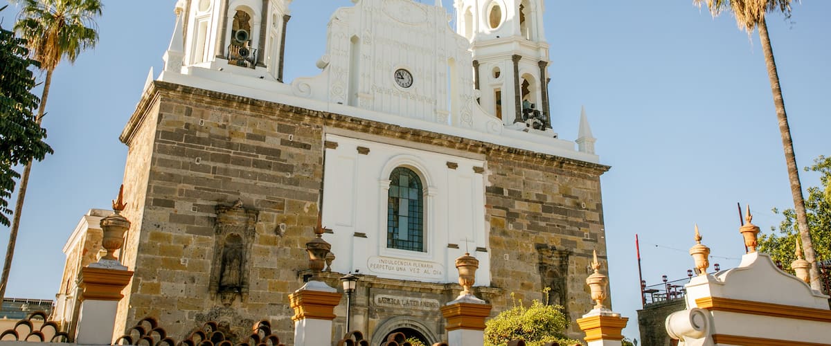 A historic white church with two bell towers and a clock, surrounded by palm trees.