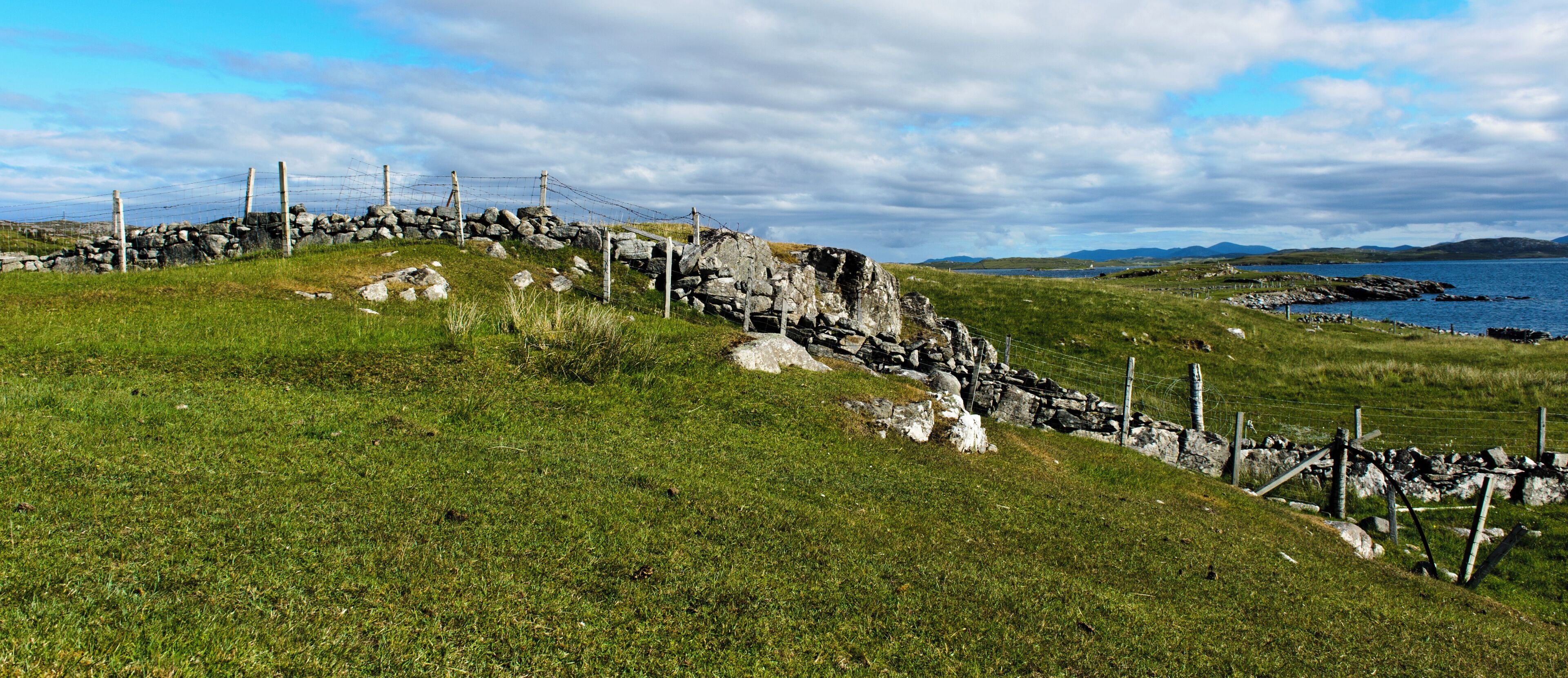 A stone wall with barbed wire, a grassy hill, and a body of water in the distance.