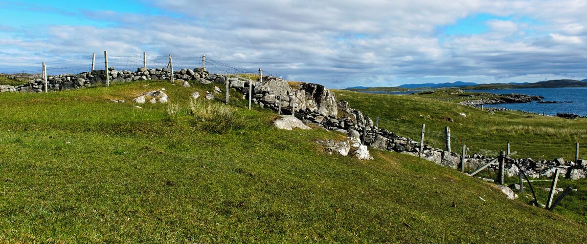 A stone wall with barbed wire, a grassy hill, and a body of water in the distance.