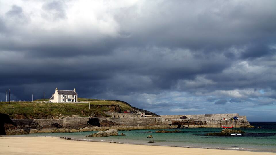 A coastal landscape with a white house on a hill, a stone wall, and the sea.