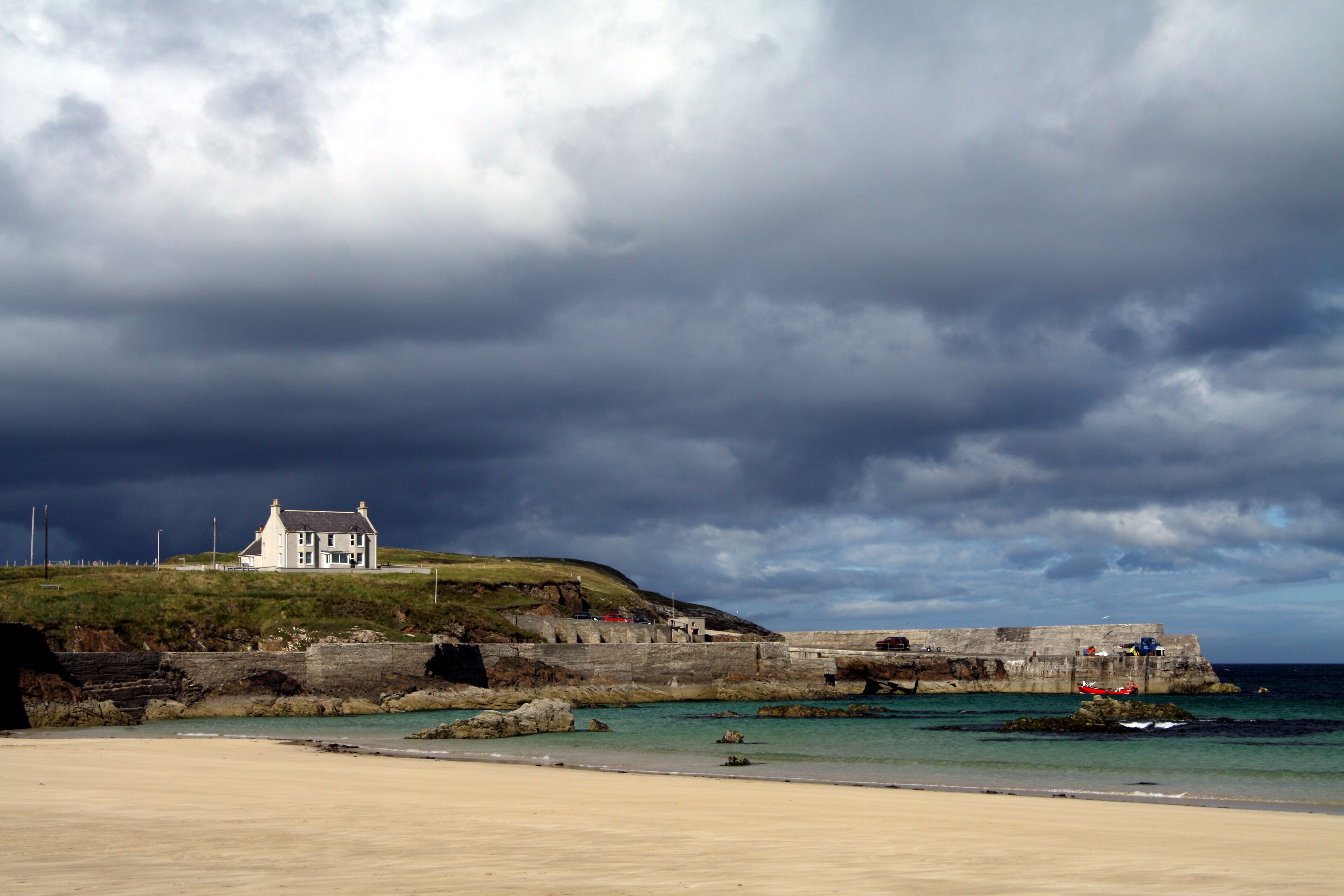 A coastal landscape with a white house on a hill, a stone wall, and the sea.