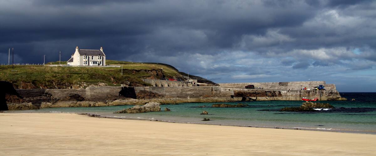 A coastal landscape with a white house on a hill, a stone wall, and the sea.
