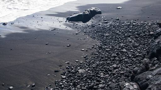 Ein Strand mit dunklem Sand, vereinzelten Kieselsteinen und einer großen Felsformation.