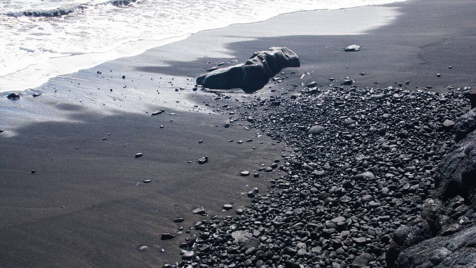 A beach with dark sand, scattered pebbles, and a large rock formation.