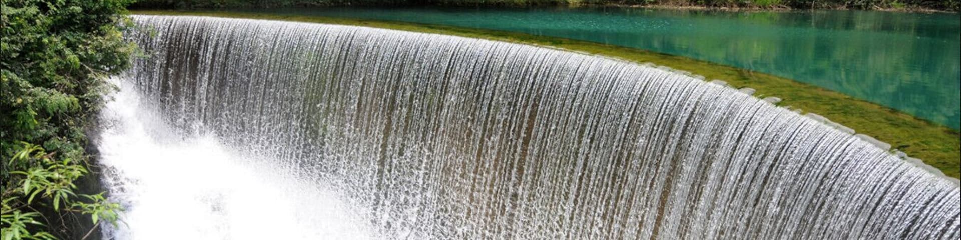 Uma cascata deságua em um lago turquesa límpido, cercado por uma floresta densa.