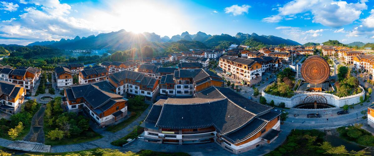 A traditional village with wooden buildings, a large circular structure, and mountains in the background.
