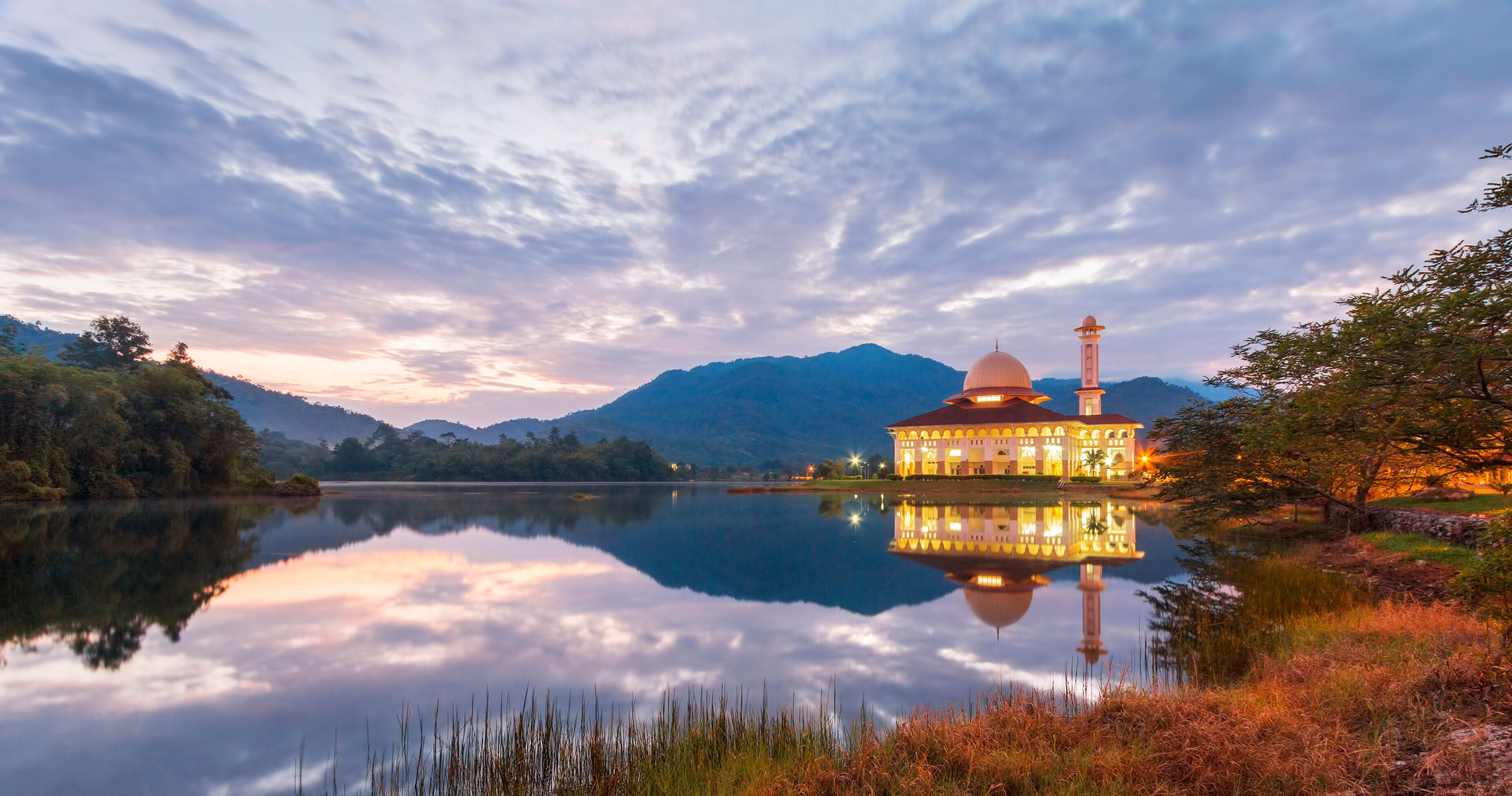 A serene lake with a reflective surface, a building with a dome and minaret, surrounded by mountains and trees.