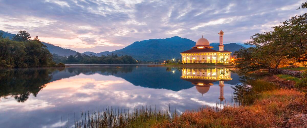 A serene lake with a reflective surface, a building with a dome and minaret, surrounded by mountains and trees.
