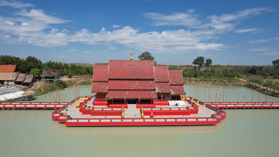 A multi-tiered temple with a red roof, situated on a floating platform in a calm body of water, surrounded by greenery and a clear sky.