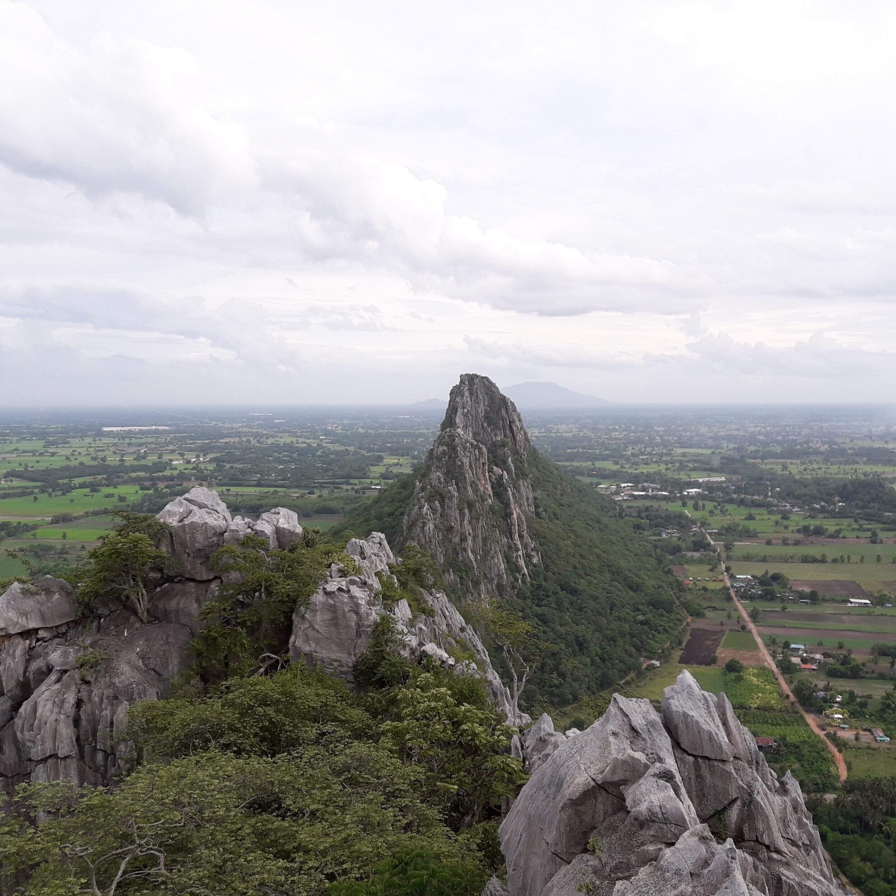 A rugged mountain peak surrounded by lush greenery and a sprawling valley with scattered buildings.