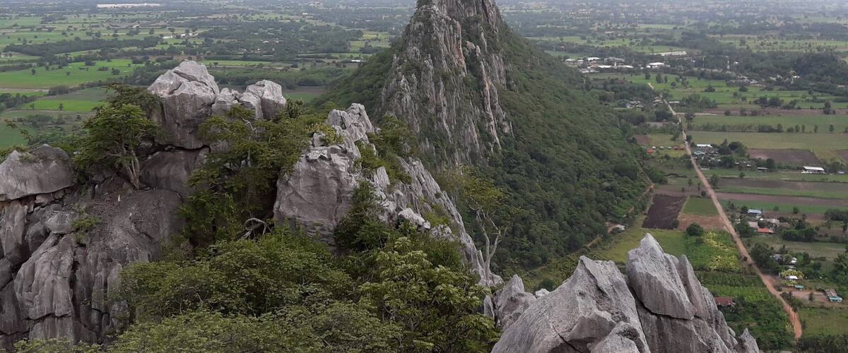A rugged mountain peak surrounded by lush greenery and a sprawling valley with scattered buildings.