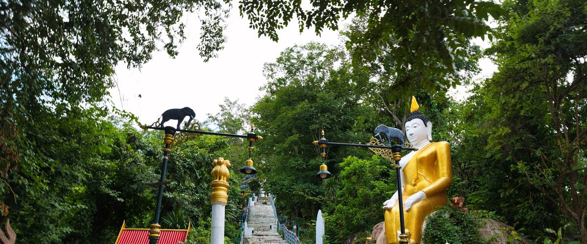 A temple with a golden statue, a white statue, and a black statue on a staircase surrounded by greenery.