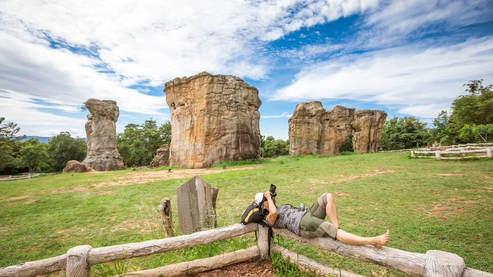 A person lying on a wooden fence in a grassy area with large rock formations in the background.