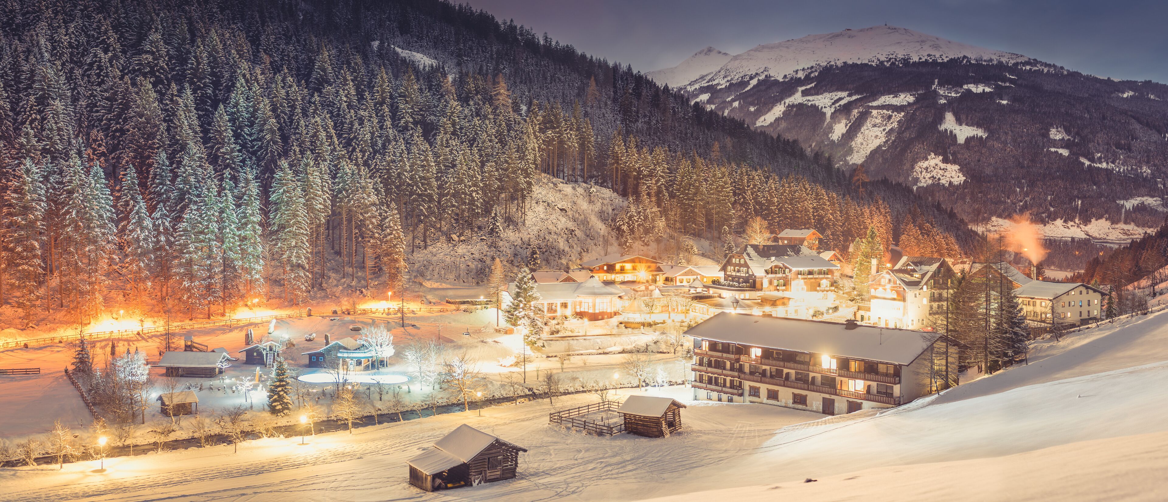 A snow-covered mountain village at night with illuminated buildings and a ski slope.