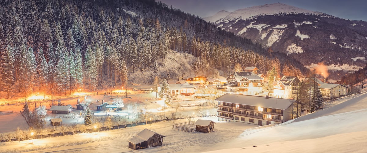 A snow-covered mountain village at night with illuminated buildings and a ski slope.