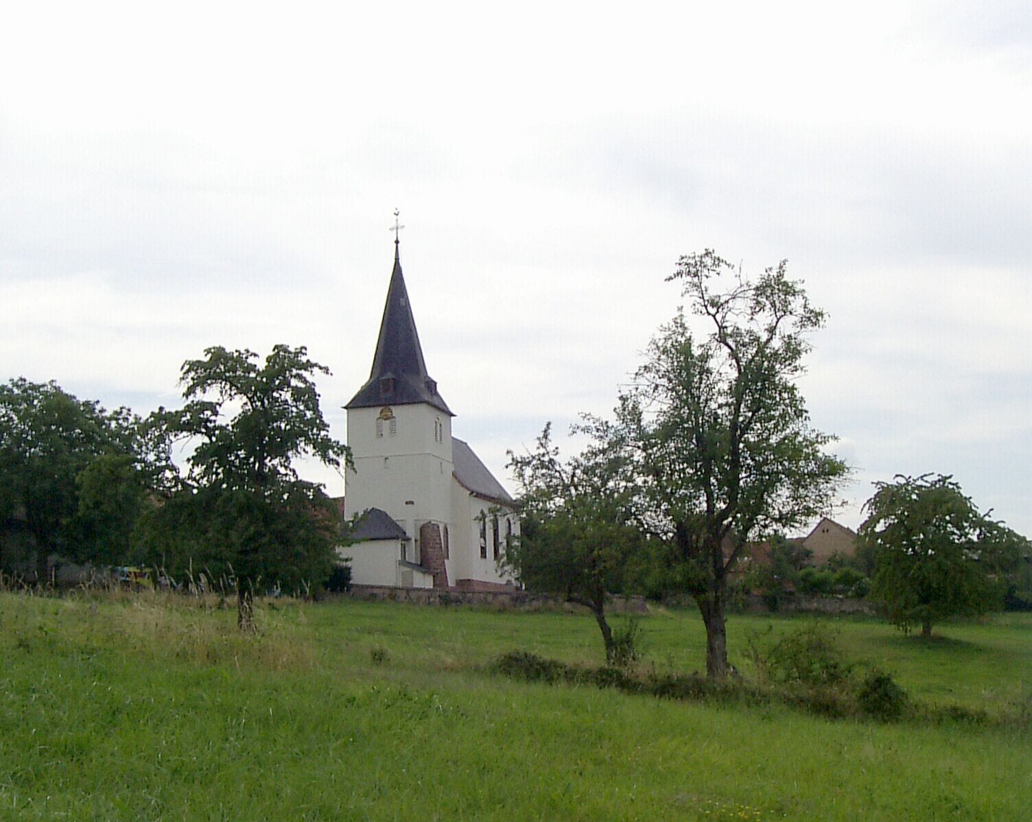 A white church with a tall steeple and a cross on top, surrounded by green grass and trees.